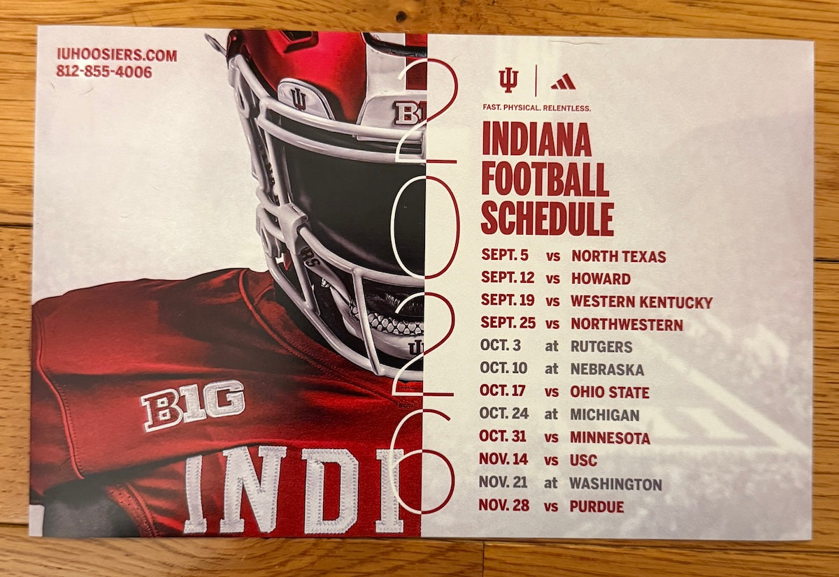 A printed football schedule poster for Indiana, laid flat on a wooden surface. The left side shows a close-up of a red and white Indiana football helmet and jersey with the word INDIA and large white numerals.