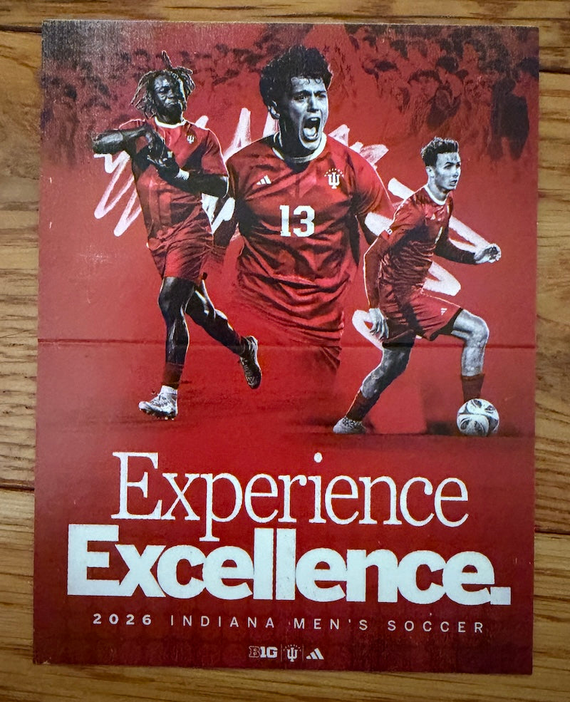A printed poster or cover design for 2026 Indiana Men's Soccer featuring three soccer players in red uniforms on a red background with the words 'Experience Excellence.' and team logos at the bottom, photographed on a wooden surface.